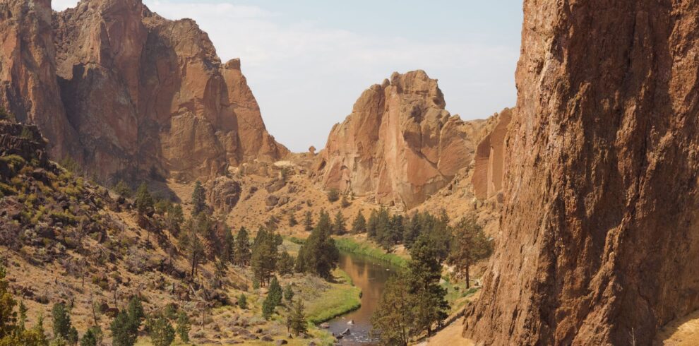 Narrow lake flowing among grassy terrain with trees surrounded with rocky cliffs against cloudy sky in national park on summer day